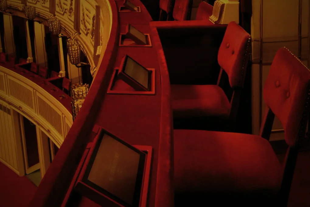 Logenbrüstung in der Wiener Staatsoper mit eingelassenen Sitzdisplays vor roten Samtstühlen.